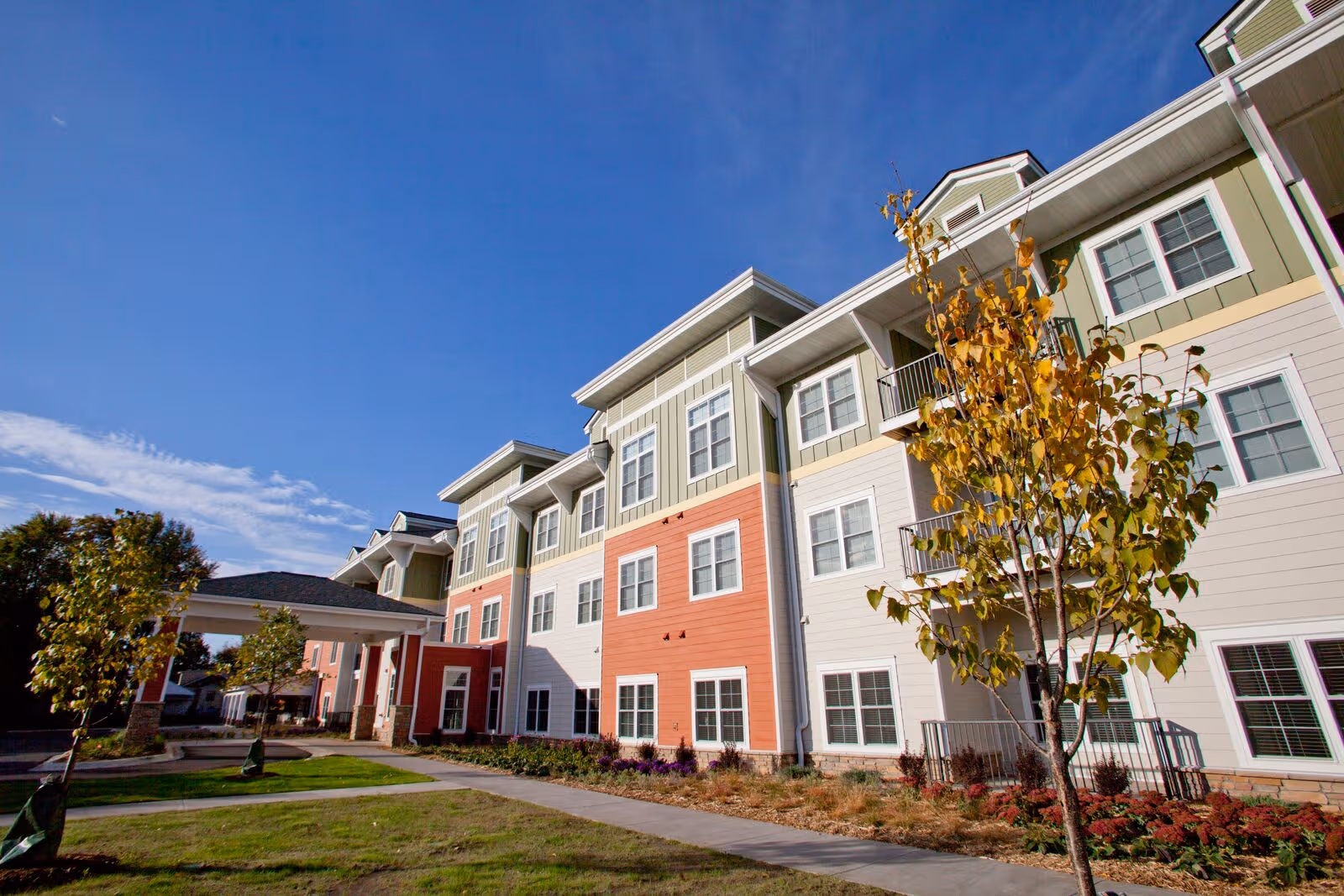 Colorful multi-story senior living building with windows, walkway, trees, and blue sky.
