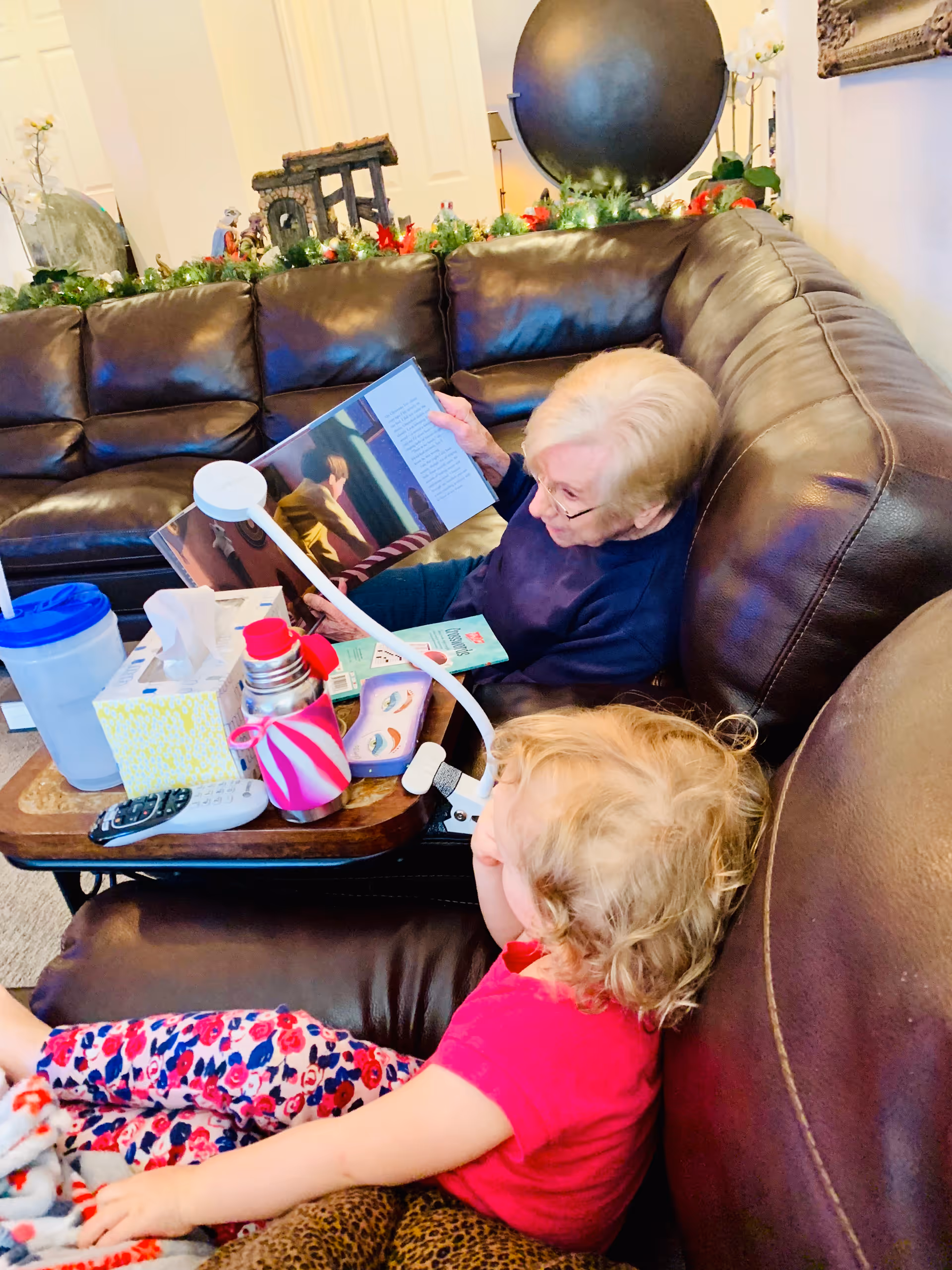 An elderly woman and a young child sitting on a large brown leather sectional sofa in a living room. The elderly woman is reading a book to the child. On a small table in front of them are a water bottle, tissue box, thermos, remote control, and other small items. The room is decorated with greenery and holiday decorations on a shelf behind the sofa.