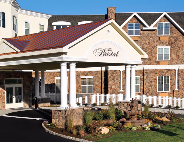 Front entrance of The Bristal assisted living building with a covered porte-cochere, white columns, stone façade and landscaped fountain.