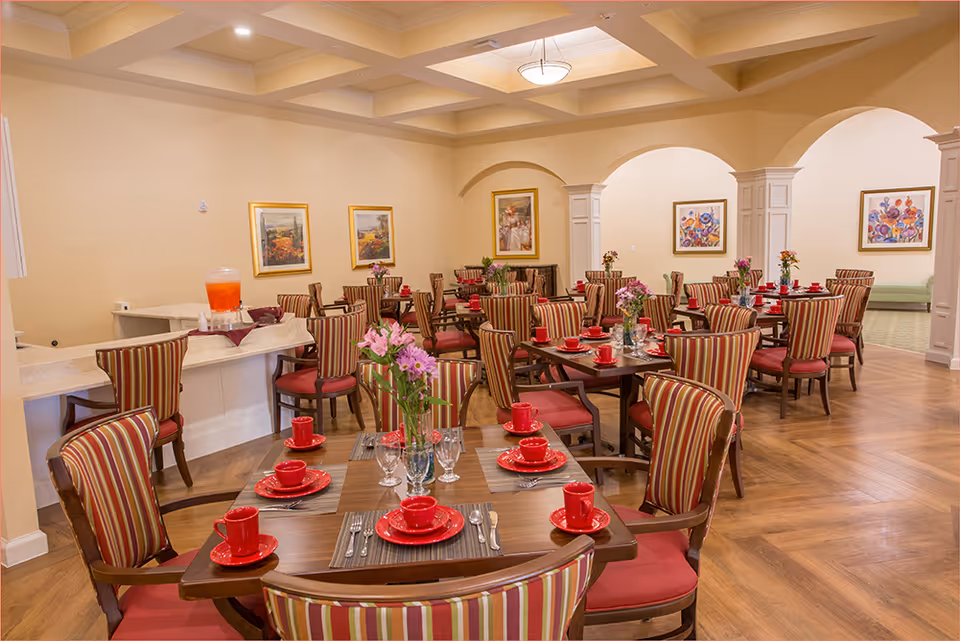 A spacious dining room with multiple wooden tables set with red plates, cups, and silverware. Each table has a vase with fresh flowers. The chairs have striped upholstery with red cushions. The room features hardwood floors, beige walls, framed artwork, and arched doorways leading to another area.
