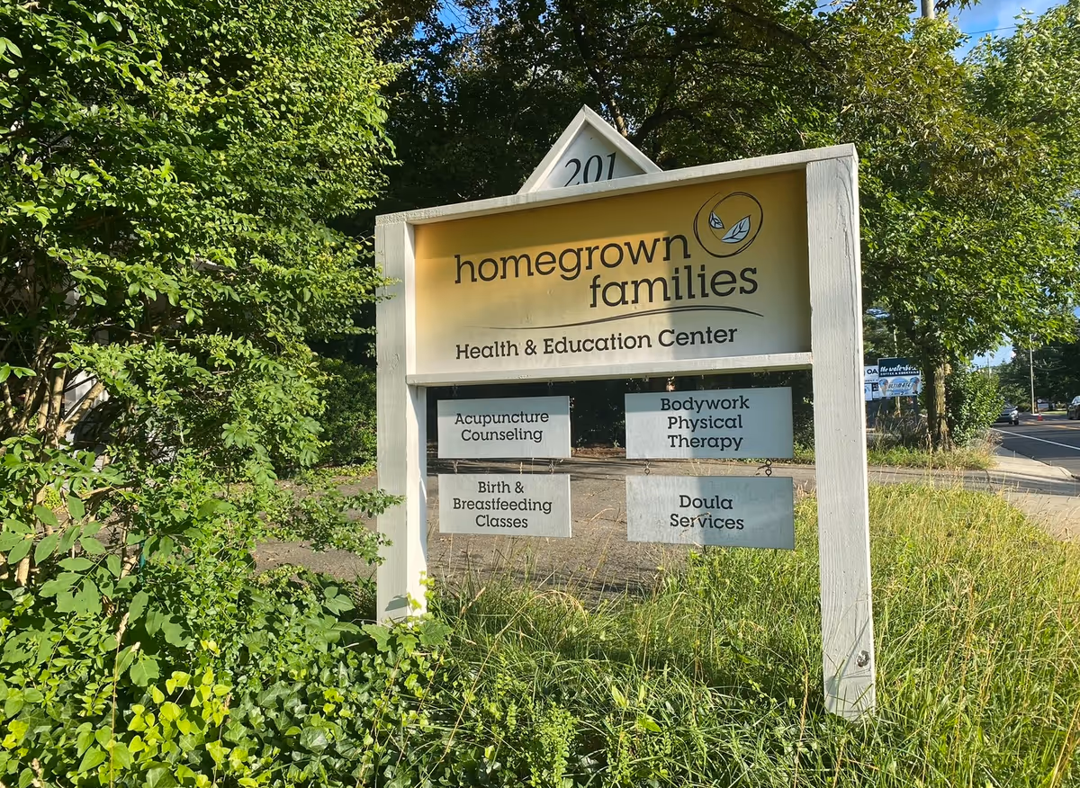 Outdoor sign for Homegrown Families Health & Education Center with service listings amid greenery.