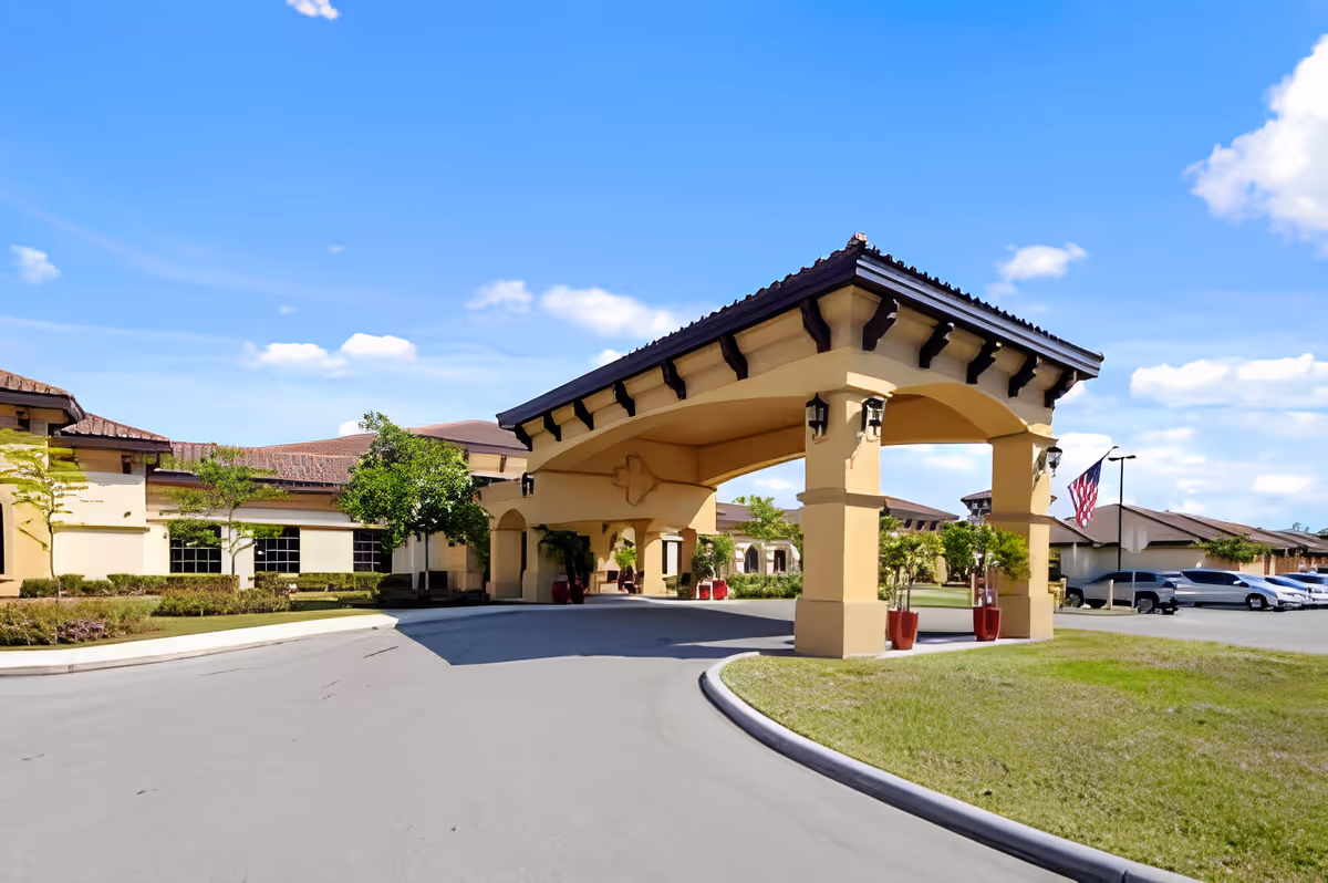 Exterior view of Discovery Commons Cypress Point facility entrance with a covered driveway, beige walls, brown tiled roof, landscaped greenery, and parked cars under a blue sky with scattered clouds.