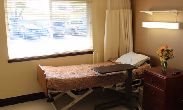 A single hospital-style bed with a brown patterned blanket and a white pillow in a room with beige walls. Next to the bed is a wooden nightstand with three drawers, a vase of flowers, and a TV remote. A window with blinds is on the left side, letting in natural light. A curtain is partially visible behind the bed.