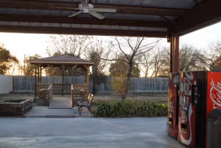 Covered outdoor pavilion area showing a wooden gazebo and bench in a yard with trees and a vending machine to the right.