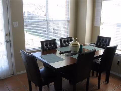 A dining area with a rectangular wooden table surrounded by six black cushioned chairs. The table has placemats and a decorative vase in the center. Large windows with blinds allow natural light to fill the room.
