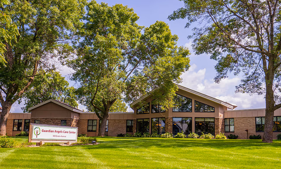 Sunny exterior view of Guardian Angels Care Center with a sign, green lawn, and trees.