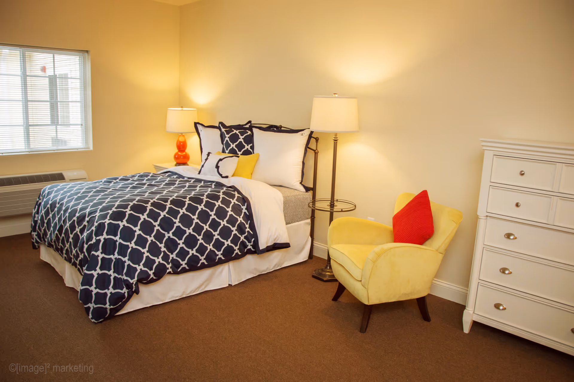 A neatly made bedroom with a navy patterned bedspread, a yellow armchair with red pillow, floor lamp, and white dresser.