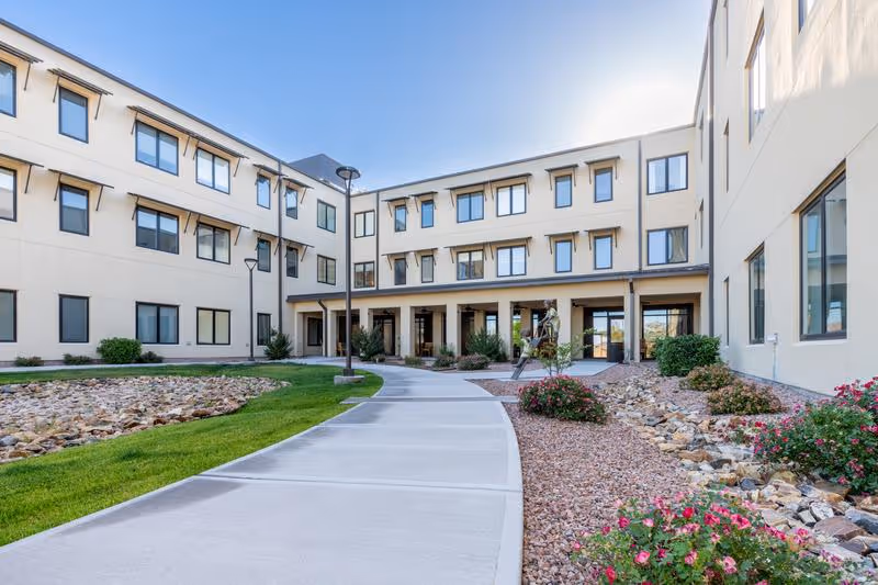 Exterior courtyard and entrance of a three-story senior living building with a paved walkway, landscaping, and multiple windows.