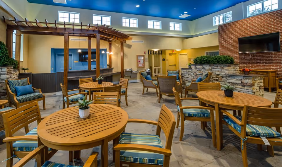 A spacious indoor common area with multiple round wooden tables and chairs with blue striped cushions. There are additional cushioned armchairs along the walls, a stone half-wall with greenery, a mounted flat-screen TV on a brick wall, and a wooden pergola structure over a counter area. The ceiling is painted blue with recessed lighting and windows near the ceiling allow natural light in.