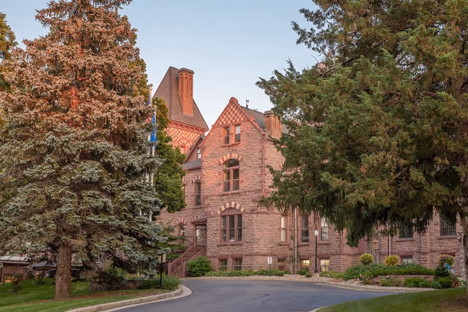 Historic stone building with steep gabled roofs and large trees framing a curved driveway.