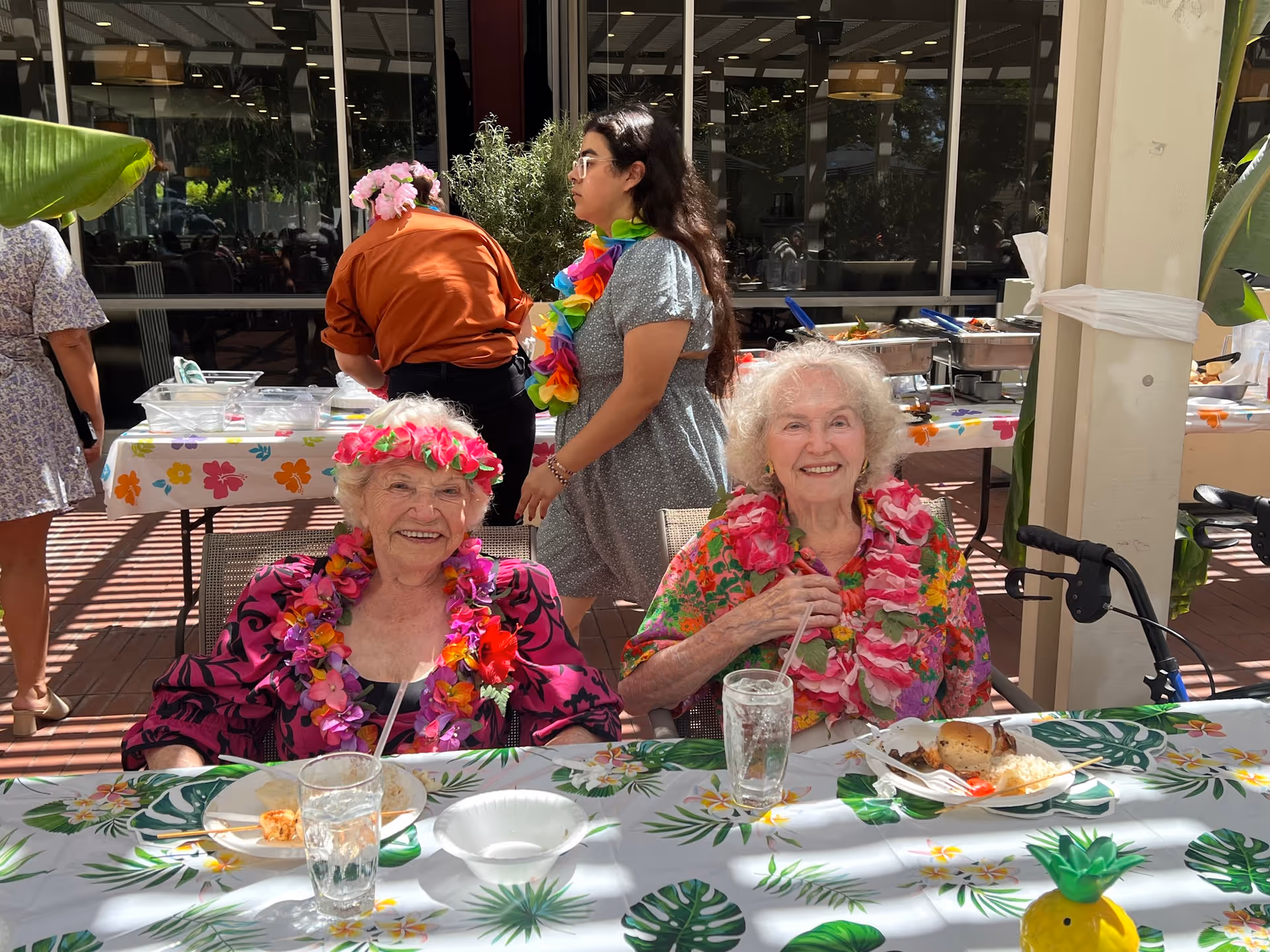 Two elderly women wearing colorful floral leis and bright clothing are seated at a table outdoors, smiling at the camera. They have plates of food and glasses of water in front of them. Behind them, a woman in a gray dress with a rainbow lei and another person in an orange shirt are near a buffet table with food trays. The setting appears festive with tropical-themed tablecloths and decorations.