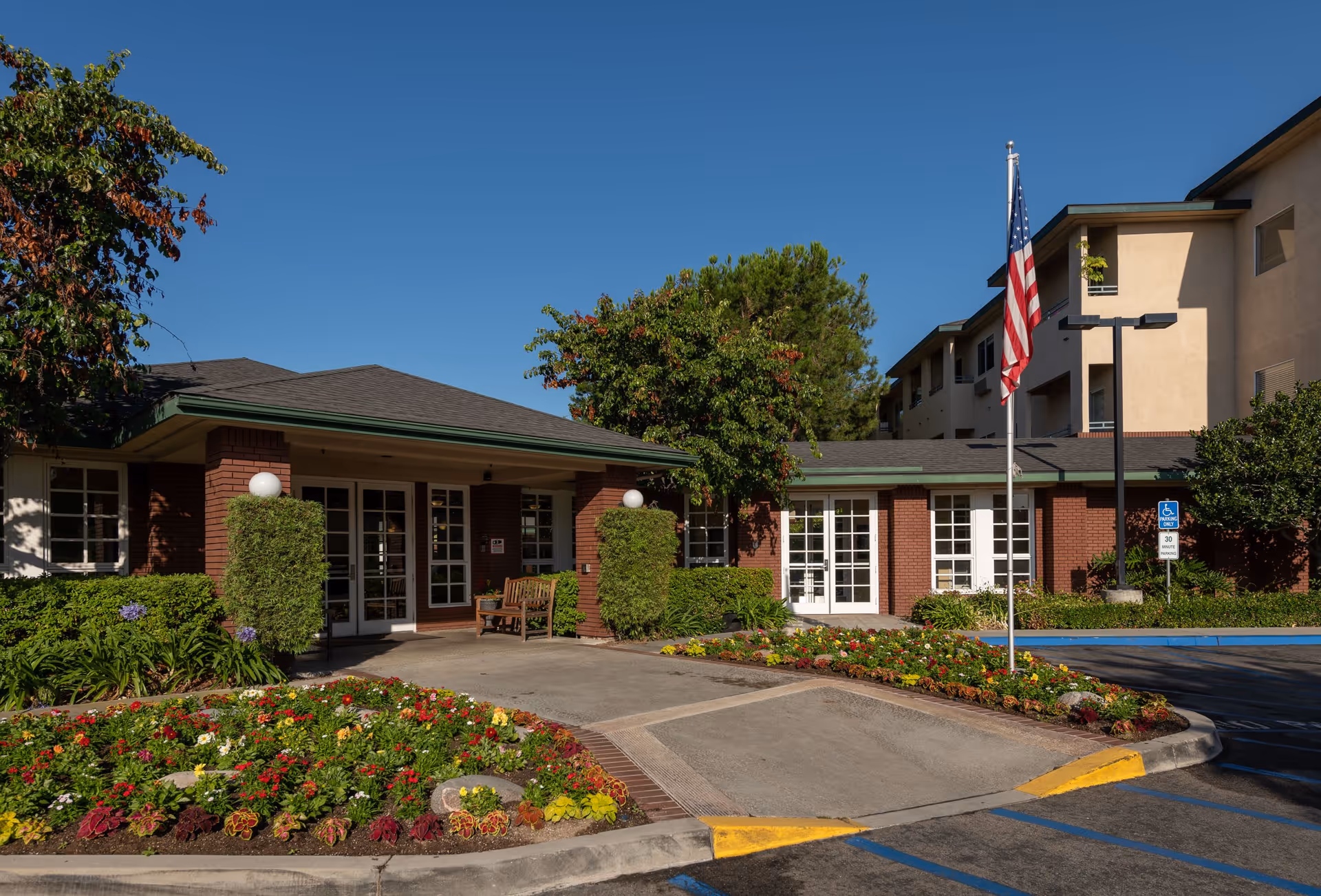 Exterior view of Carmel Village senior living facility showing a brick building with large windows and a green roof. There is a well-maintained flower bed with colorful flowers in front, a flagpole with an American flag, and a parking area with a handicapped parking sign. The sky is clear and blue.