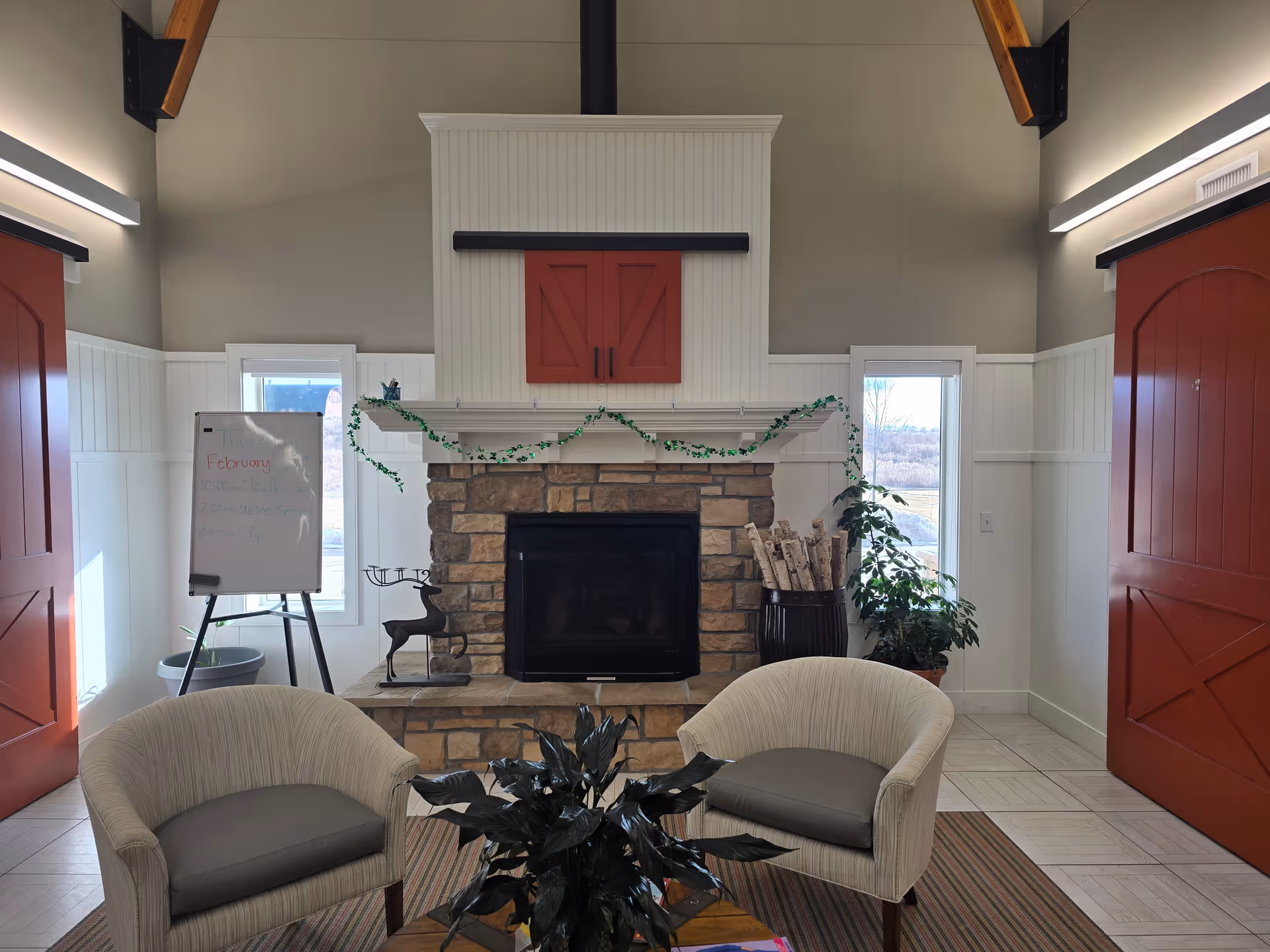 Cozy living room with stone fireplace, red doors, garland, two beige chairs, and potted plants.