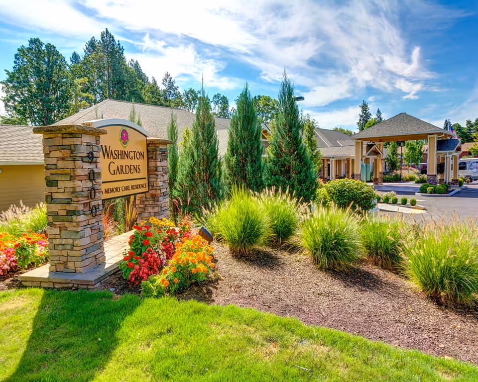 Stone sign reading 'Washington Gardens Memory Care' at the landscaped front entrance with flowers, shrubs, and the facility's covered porte-cochère.