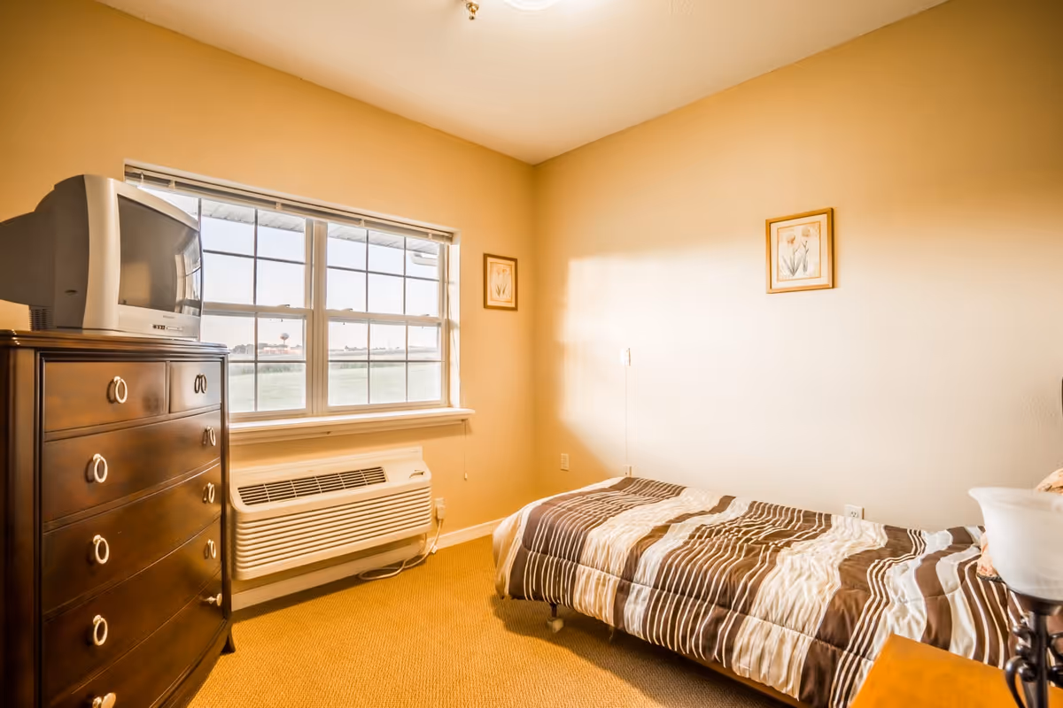A small bedroom with a single bed covered in a brown and beige striped comforter. There is a wooden dresser with a CRT television on top, a window with a view of the outdoors, and two framed pictures hanging on the beige walls. The room has a beige carpet and a wall-mounted air conditioning unit below the window.
