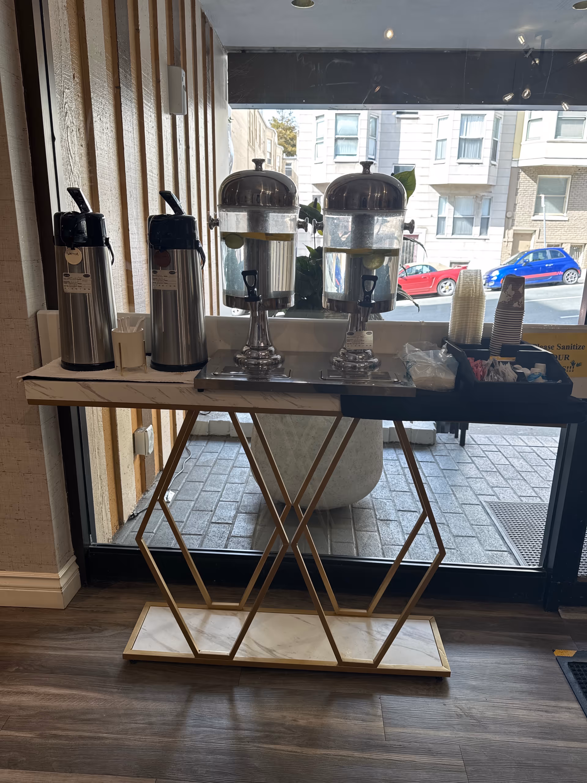 Self-serve coffee station with two urns and supplies beside a glass storefront window.