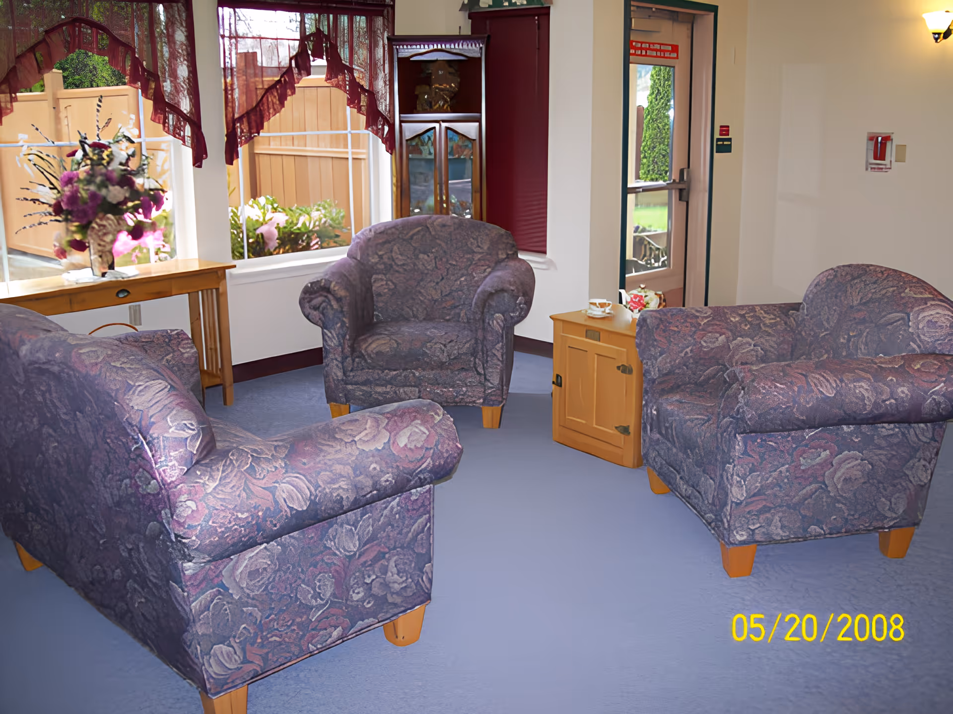 A cozy seating area with three floral-patterned armchairs arranged around a small wooden cabinet with a teacup on top. The room has light blue carpet, a wooden side table with a flower arrangement near a window with maroon lace curtains, and a glass door leading outside. The walls are light-colored with a wall sconce and a fire alarm visible.