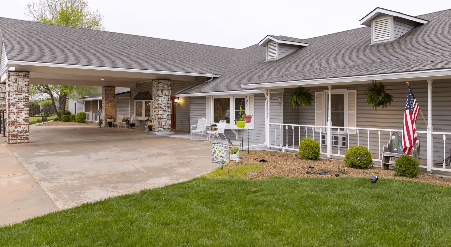 Exterior view of Homestead Assisted Living of Derby showing a covered driveway entrance supported by brick pillars, a gray building with white trim, a porch with white railings, hanging plants, an American flag, and green lawn in the foreground.