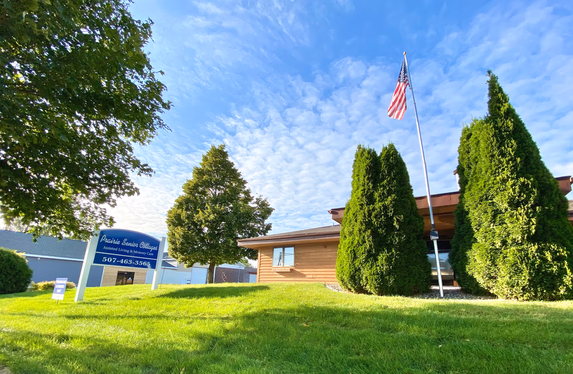 Front view of Prairie Senior Cottages building with sign, flag, and green lawn.
