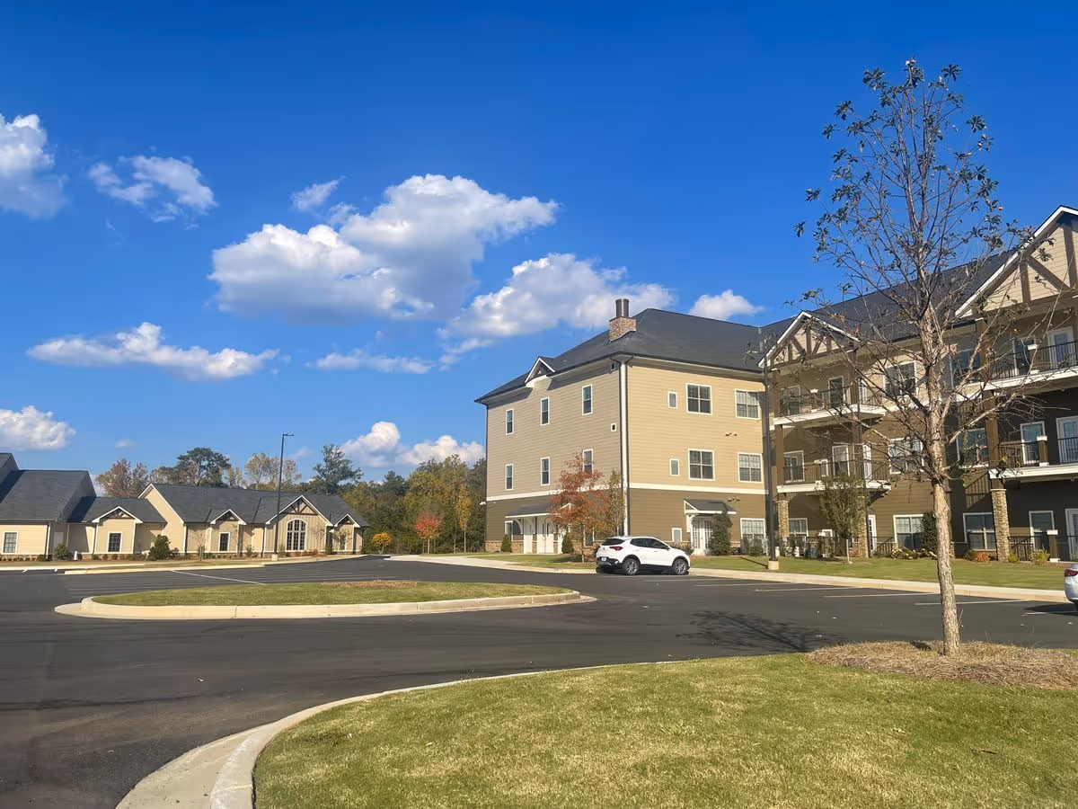 Front exterior of a multi-story senior living building with a parking lot, smaller single-story units to the left, and a clear blue sky.