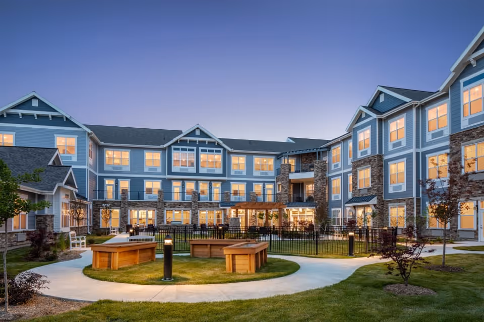 Exterior view of a senior living facility named StoryPoint Northville during dusk, showing a three-story building with many lit windows, a well-maintained lawn, paved walkways, wooden benches, small trees, and outdoor lighting.