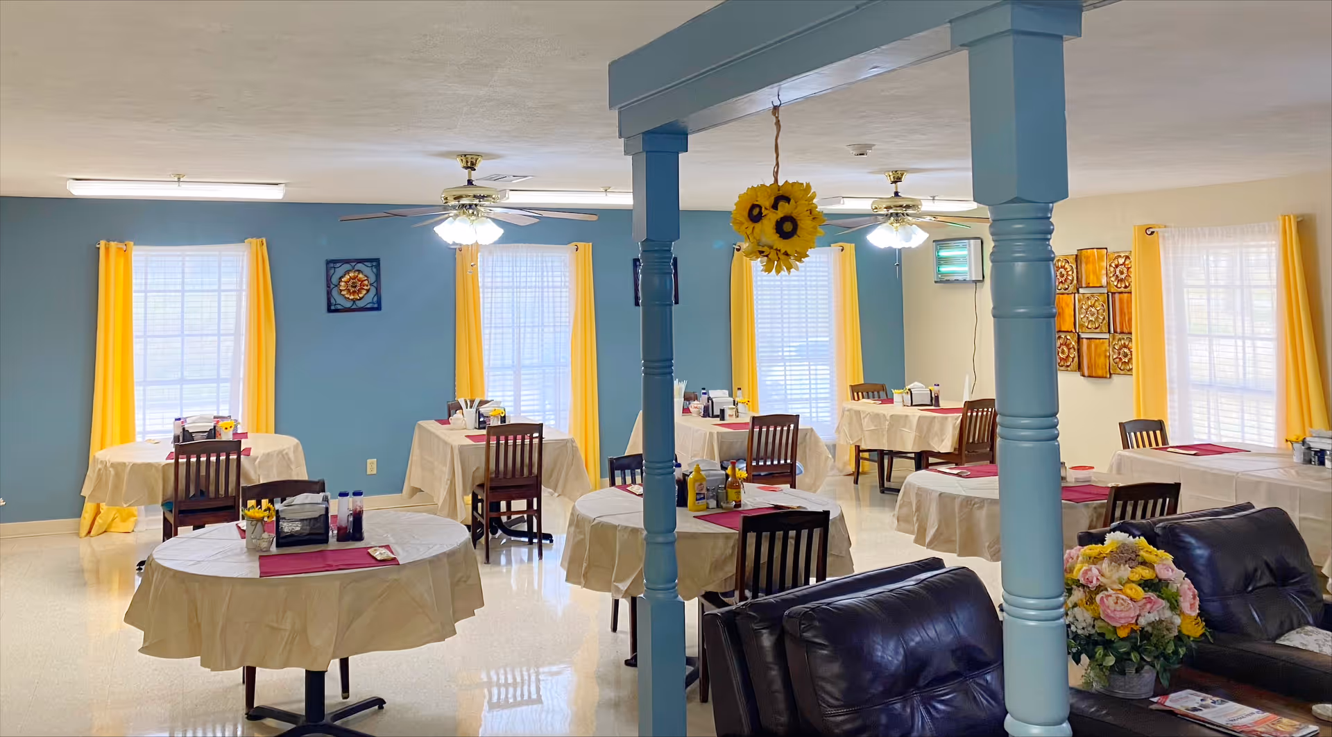 Bright dining room with round tables draped in beige cloths, wooden chairs, blue walls, yellow curtains and sunflower decorations.