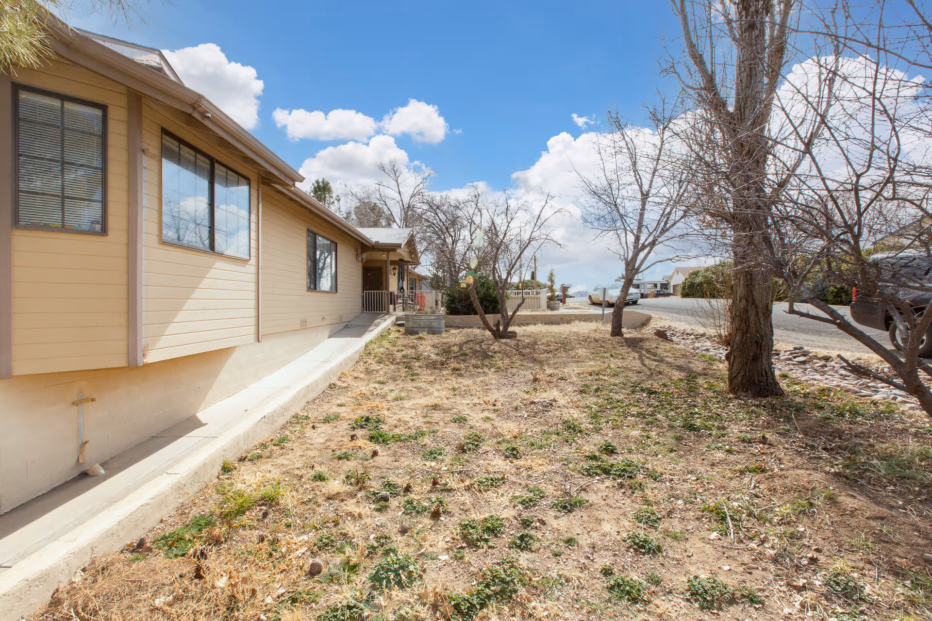 Exterior view of a beige single-story building with several windows, a small porch, and a dry, patchy yard with sparse vegetation and leafless trees under a partly cloudy blue sky.