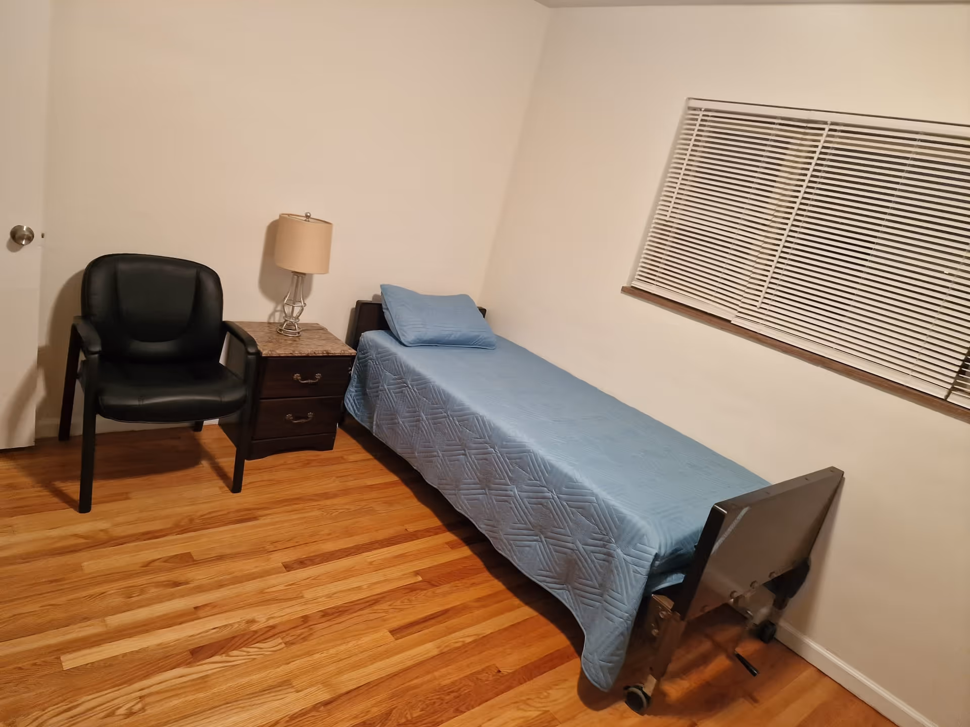 A simple bedroom with a single bed covered in a blue quilt and matching pillow, a wooden nightstand with a lamp, a black armchair, and a window with closed blinds. The floor is wooden and the walls are plain white.