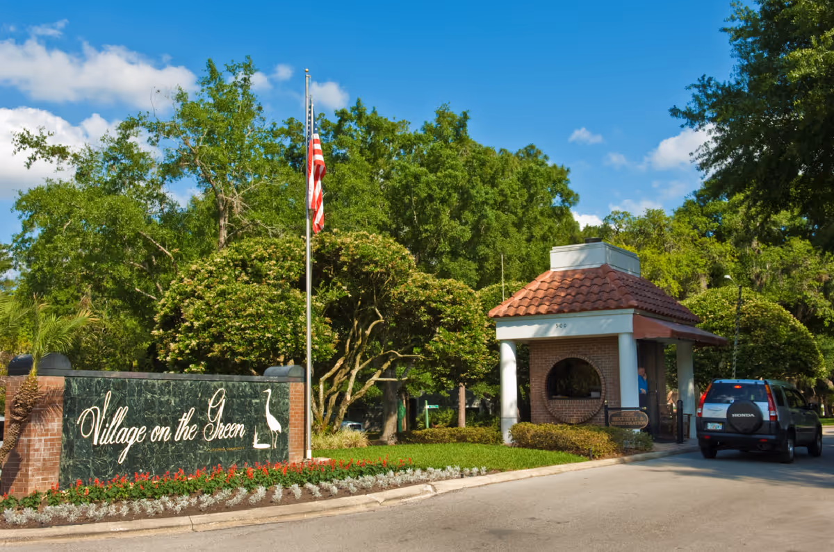 Entrance to the Village on the Green community showing the sign, flagpole, guardhouse and a car at the driveway.