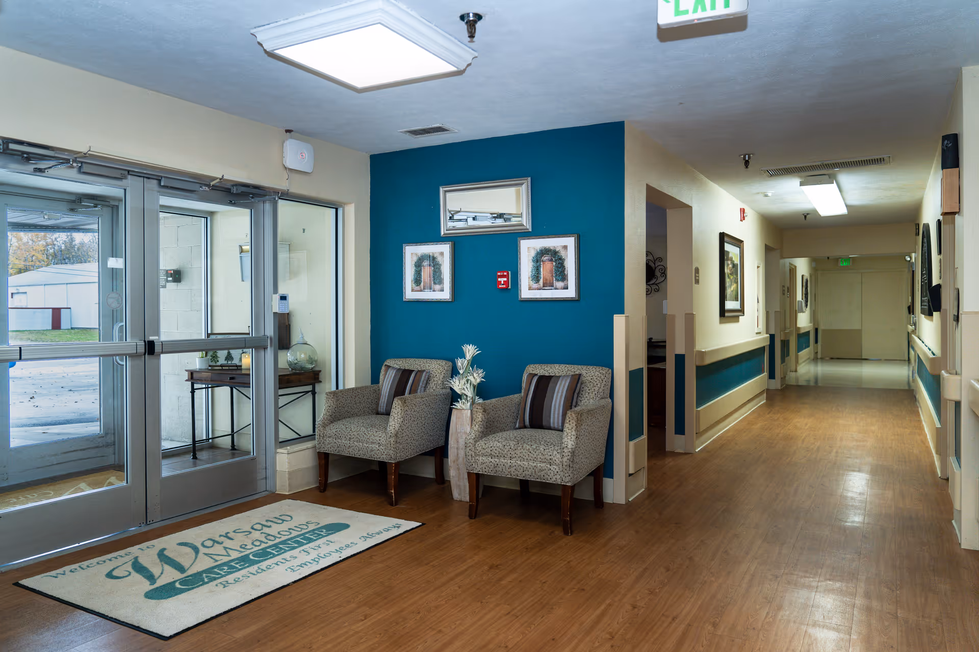 Entrance lobby with glass double doors, two armchairs by a teal accent wall and a long hallway leading inward.