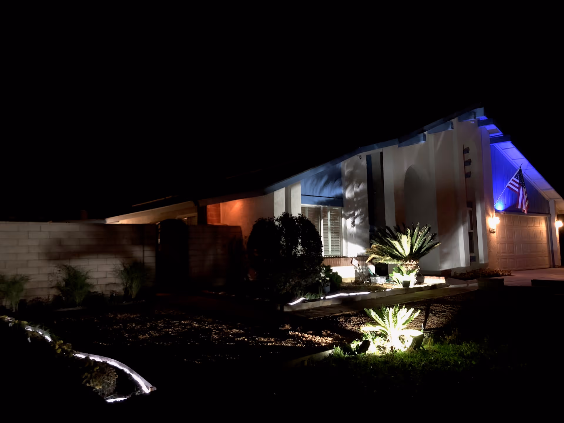 Night view of a residential building with exterior lighting illuminating plants and the front facade. The building has a garage with an American flag displayed above it and a blue light highlighting the upper part of the structure.