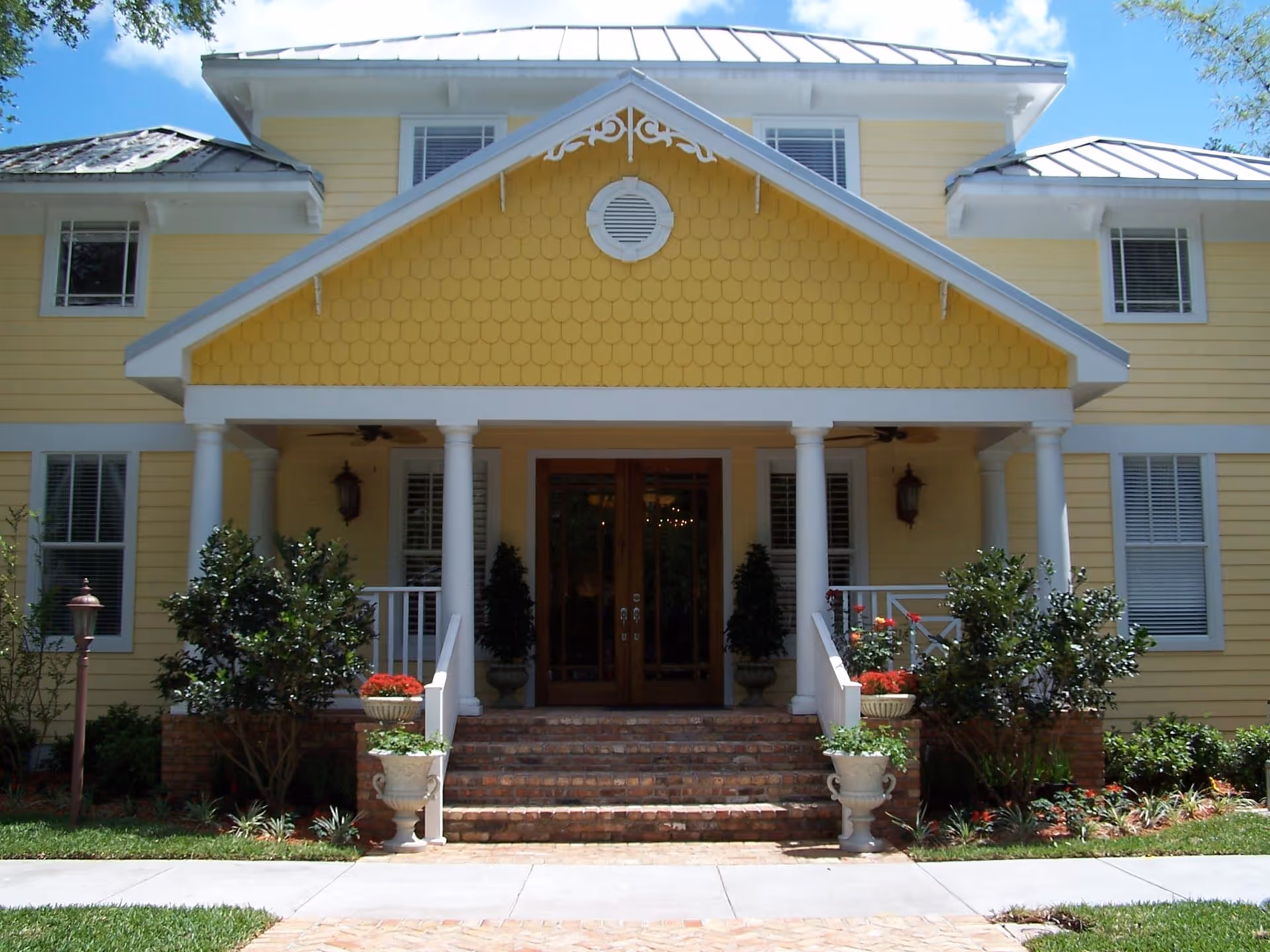 Front exterior view of a yellow two-story building with white trim, a covered porch supported by white columns, brick steps leading to double wooden doors, and landscaping with shrubs and potted plants.