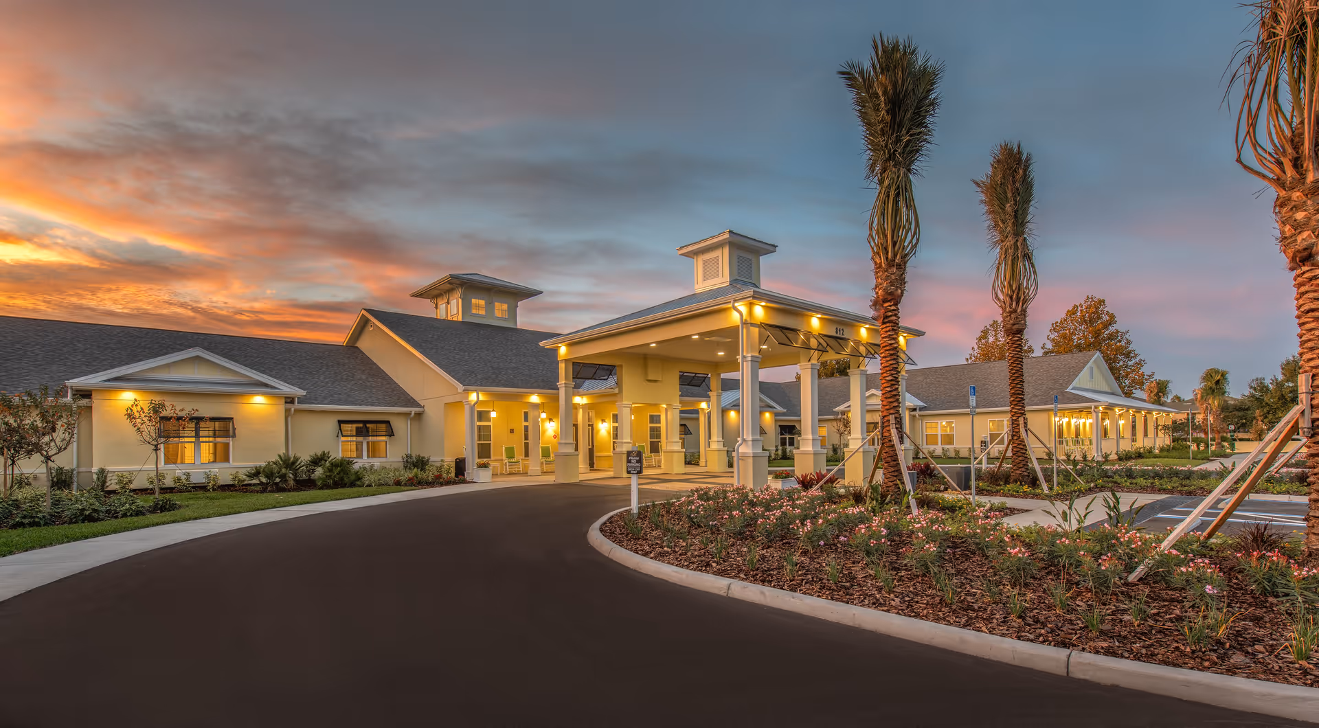Sunset exterior view of The Addison of Port Orange facility with entrance, palms, and landscaped drive.