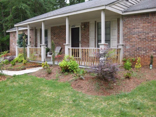 Front exterior of a single-story brick building with a covered porch, railing, chairs, and a landscaped lawn.