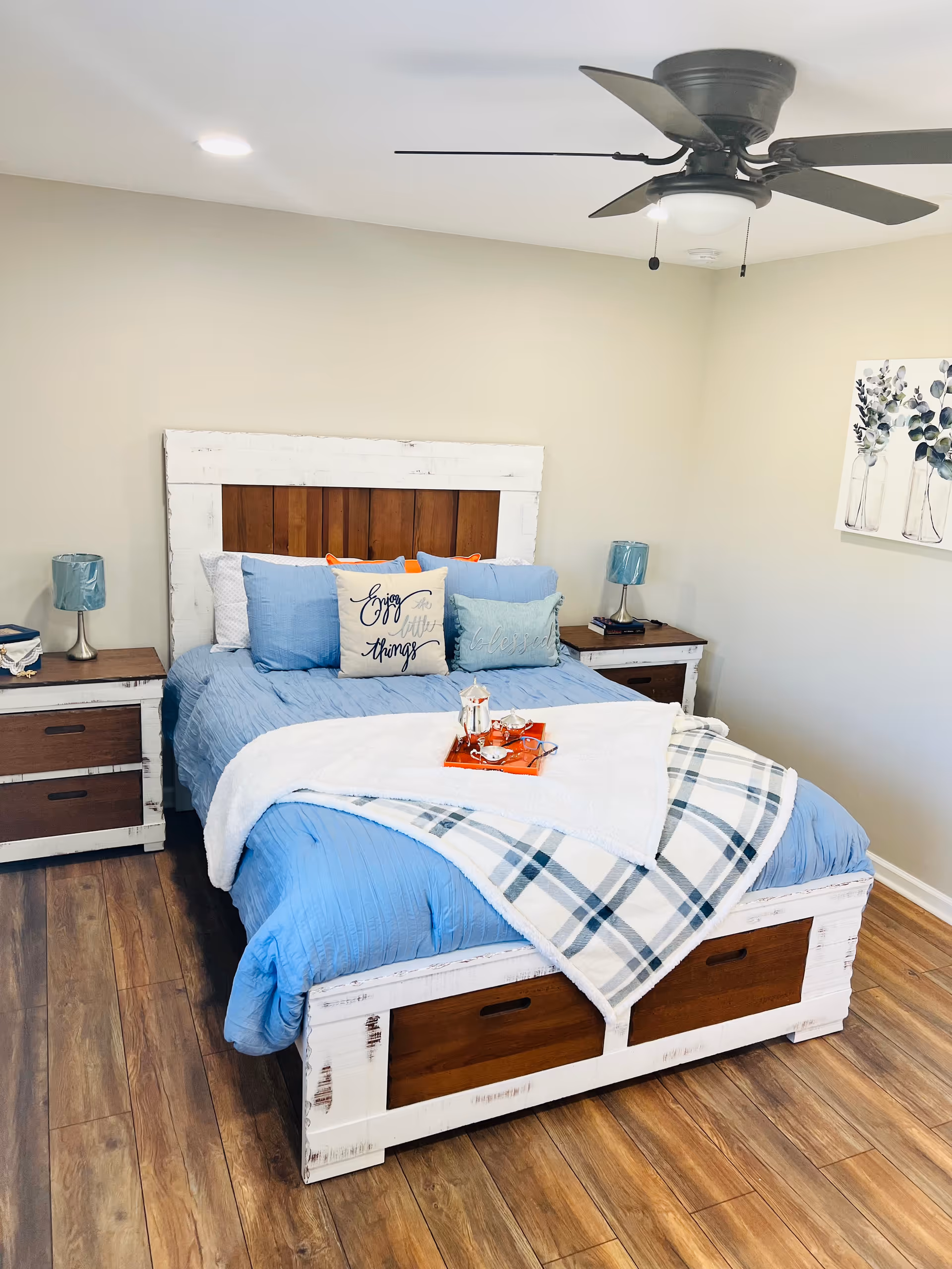 Bright bedroom with a distressed white bed frame, blue bedding, nightstands, and ceiling fan.