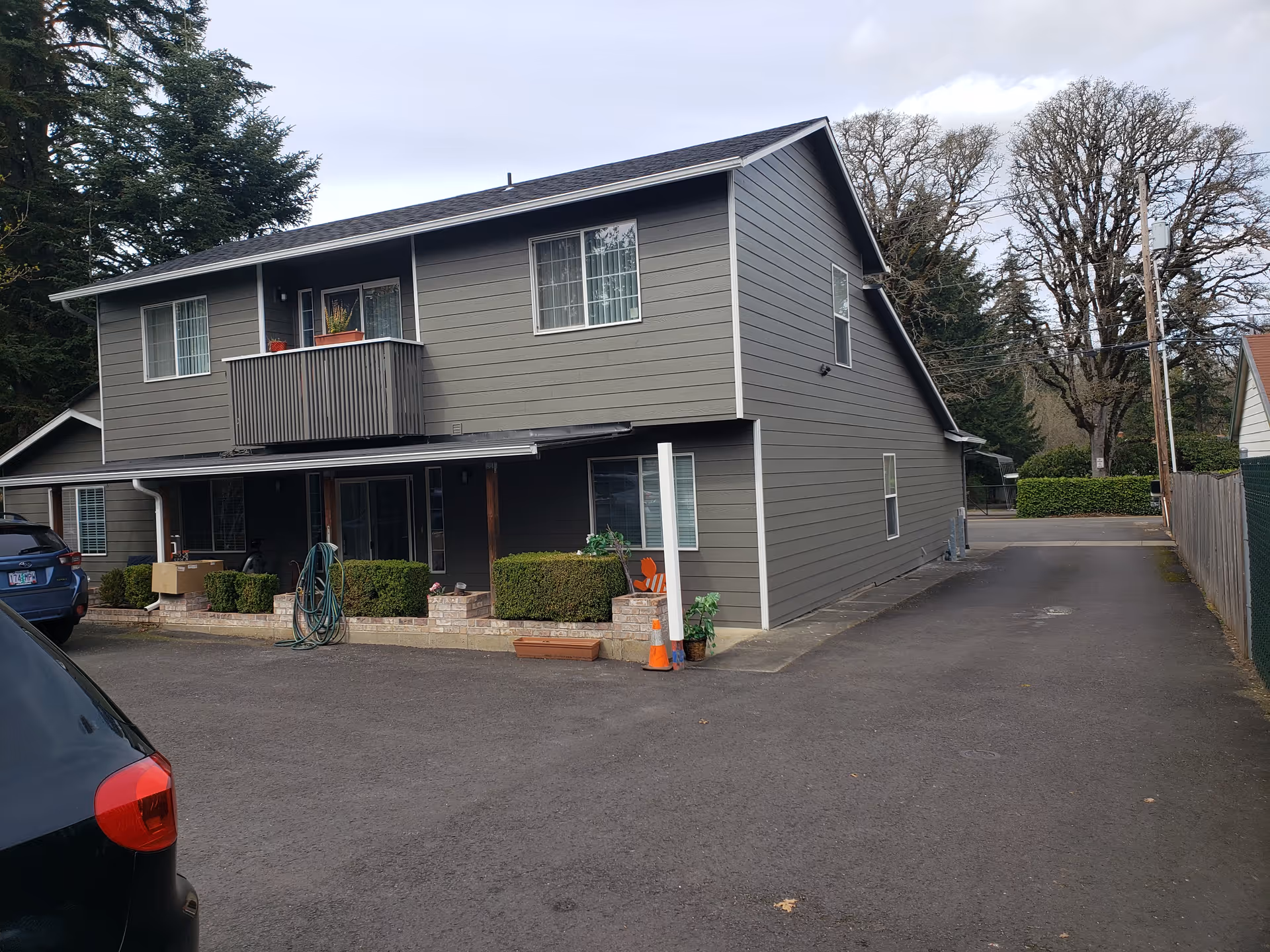 Two-story gray building with a driveway, hedges, and parked cars in front.