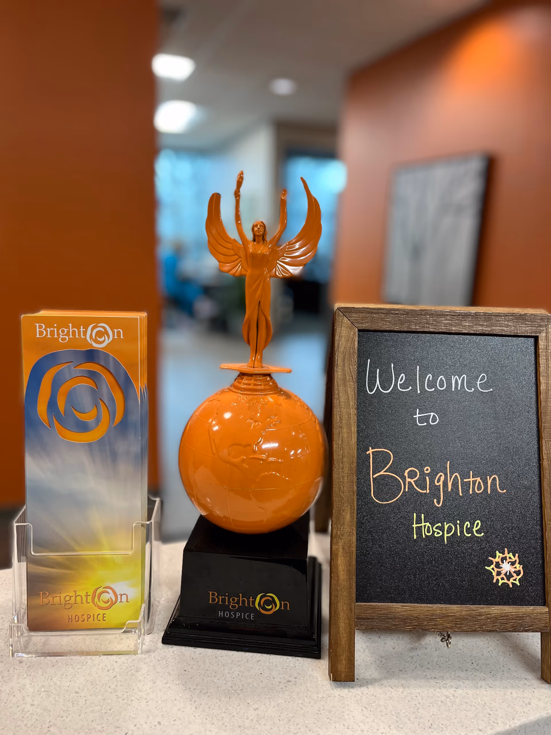 Interior reception area with Brighton Hospice branding, chalkboard welcome sign, and orange statue centerpiece.