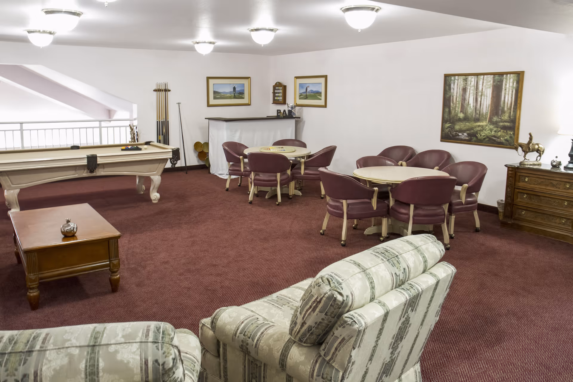A recreational room with a pool table, two round tables each surrounded by four purple chairs, a small bar area with stools, a wooden coffee table, and two patterned armchairs. The room has maroon carpet, white walls with framed paintings, and ceiling lights.