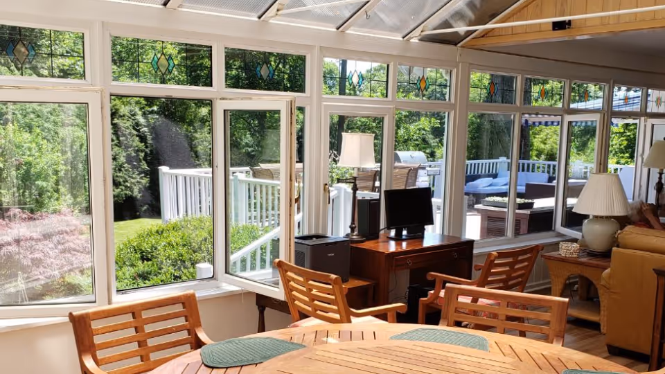 Sunlit sunroom with large windows, wooden dining table, and chairs overlooking outdoor patio.