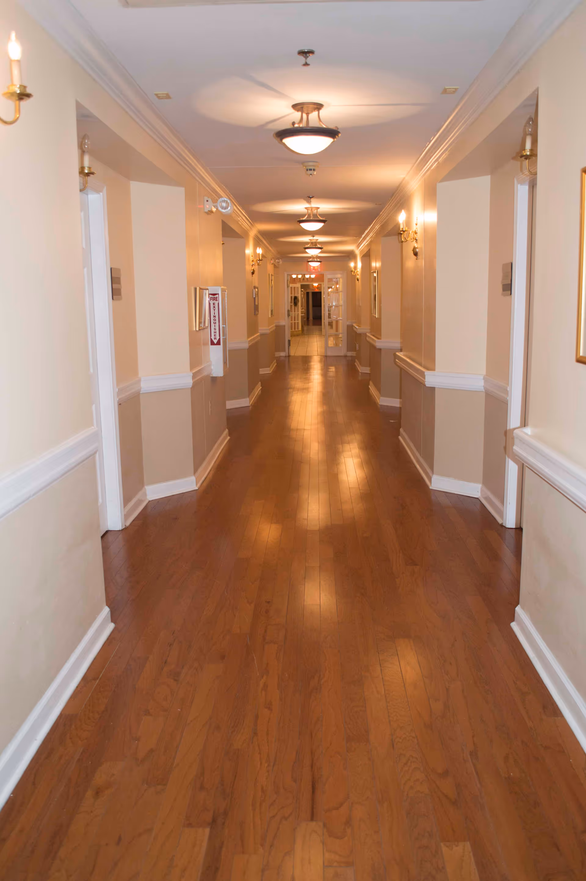 A long, well-lit hallway with wooden flooring and beige walls featuring white trim. The hallway has multiple doorways on both sides, wall-mounted light fixtures, and ceiling lights. At the end of the hallway, there is a glass door with an exit sign above it.