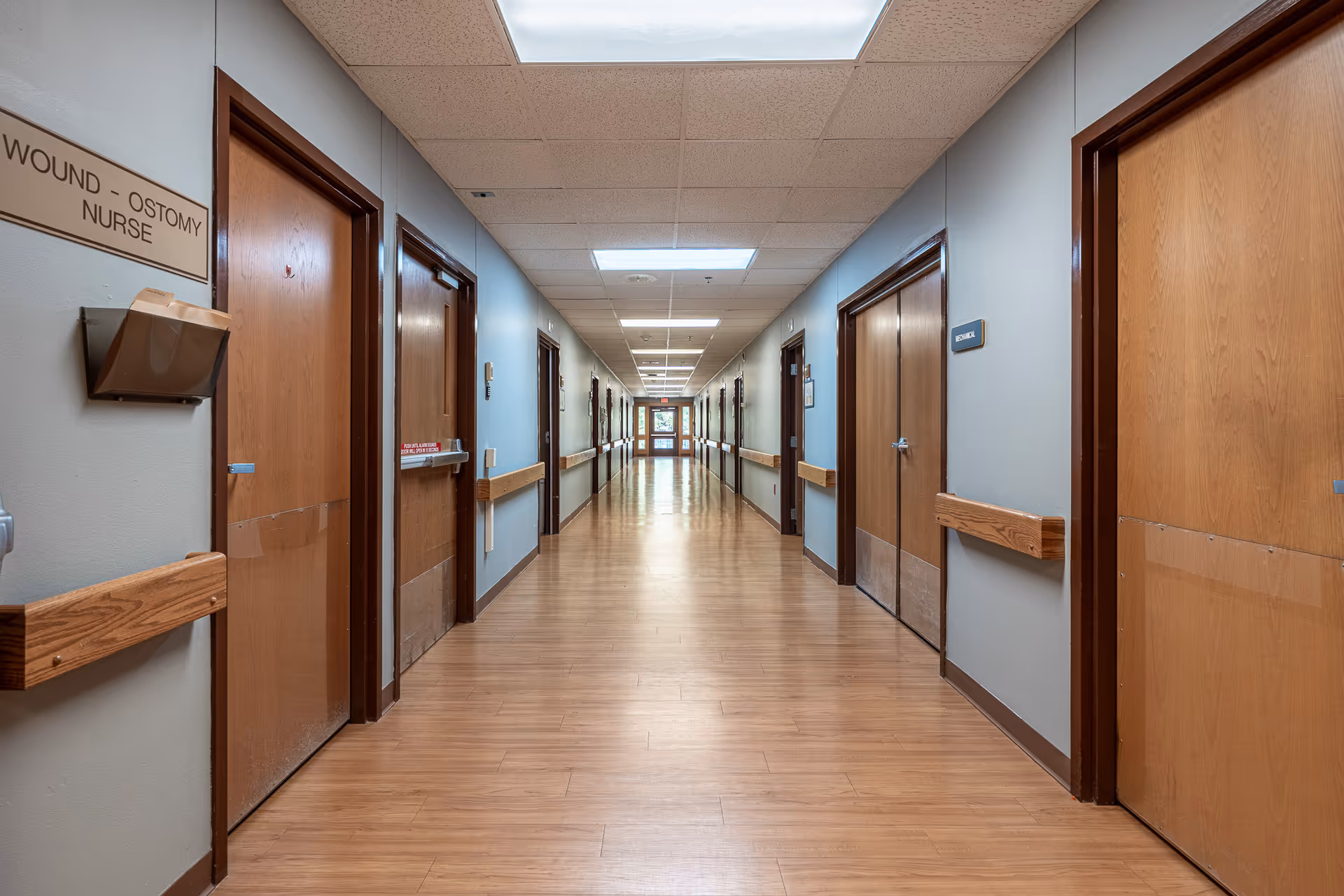 Long interior corridor with wooden doors, wall handrails, and bright ceiling lights.