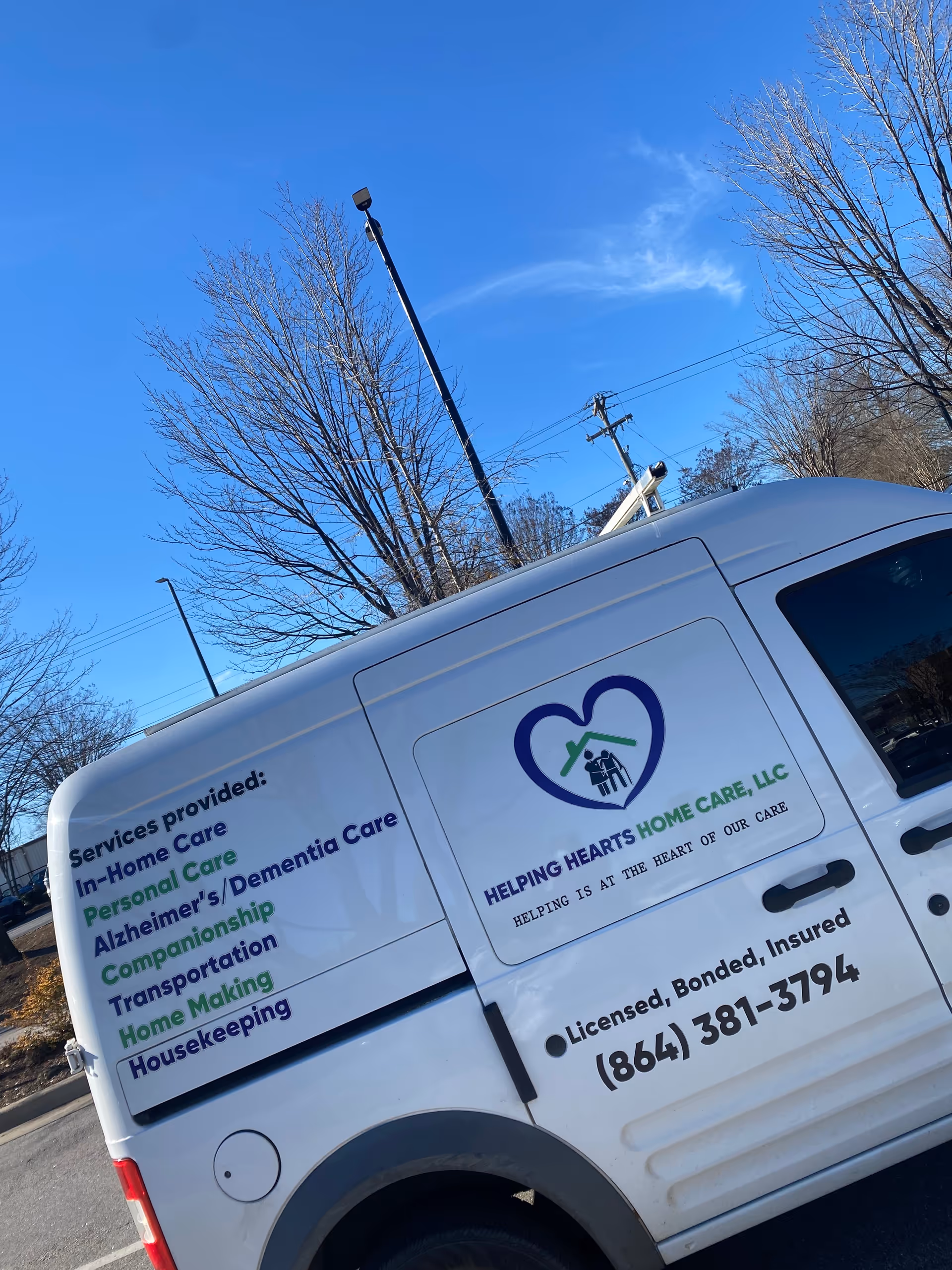 White van for Helping Hearts Home Care LLC with logo parked outdoors near bare trees.
