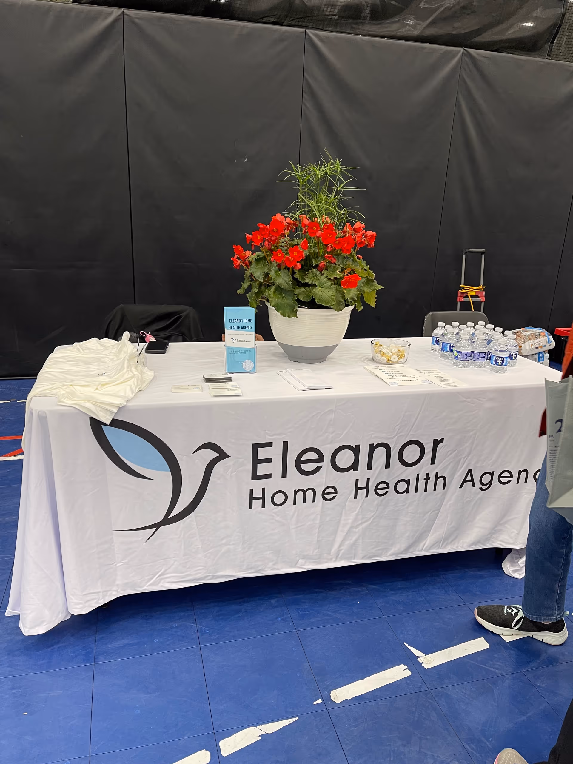 Table with Eleanor Home Health Agency banner, large flower pot centerpiece, and bottled water on display.