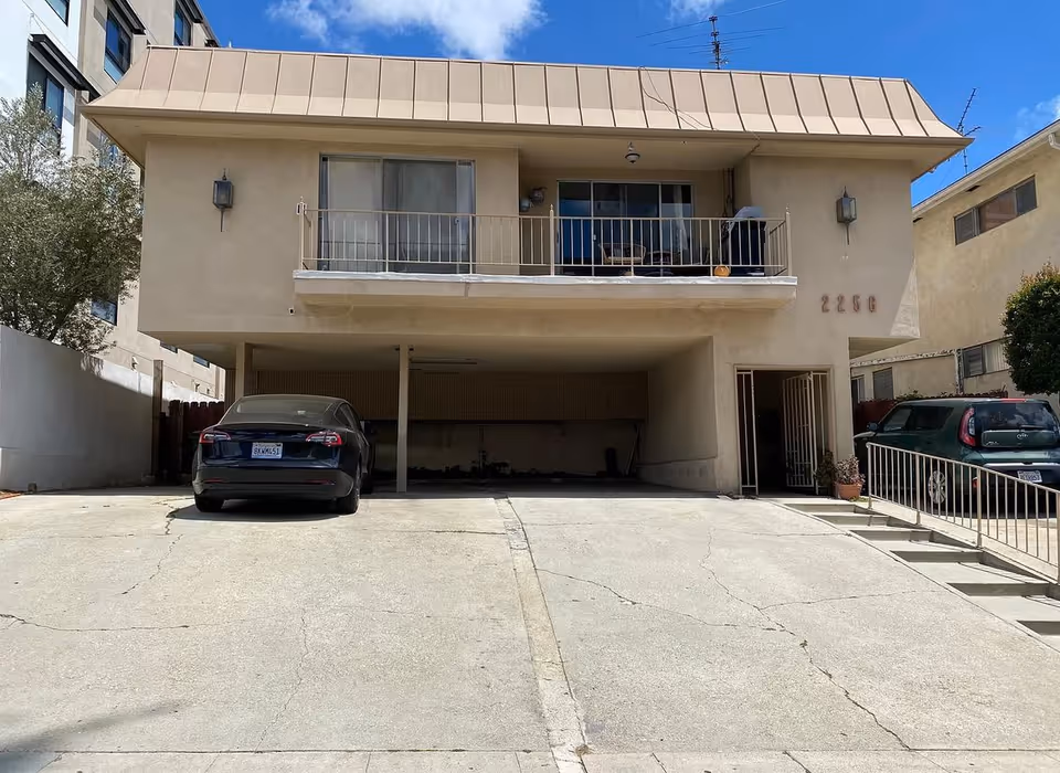 Two-story beige building with a balcony and parked cars under a carport.