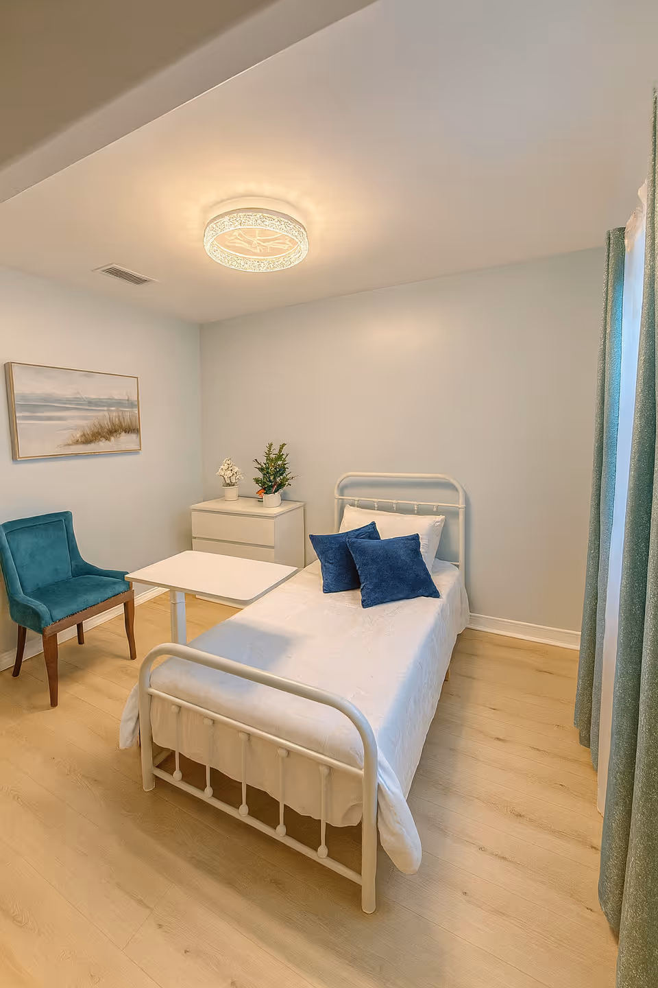 Single-bedroom in a senior living facility with light blue walls, white furniture, and blue cushions.