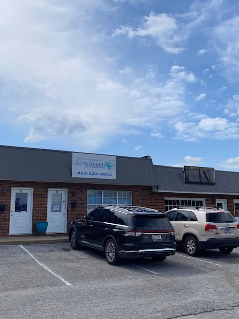 Brick building with a Visiting Angels sign, two parked cars, and a clear blue sky.