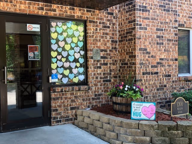Brick building exterior with front door, window decorated with hearts, and a small garden bed.