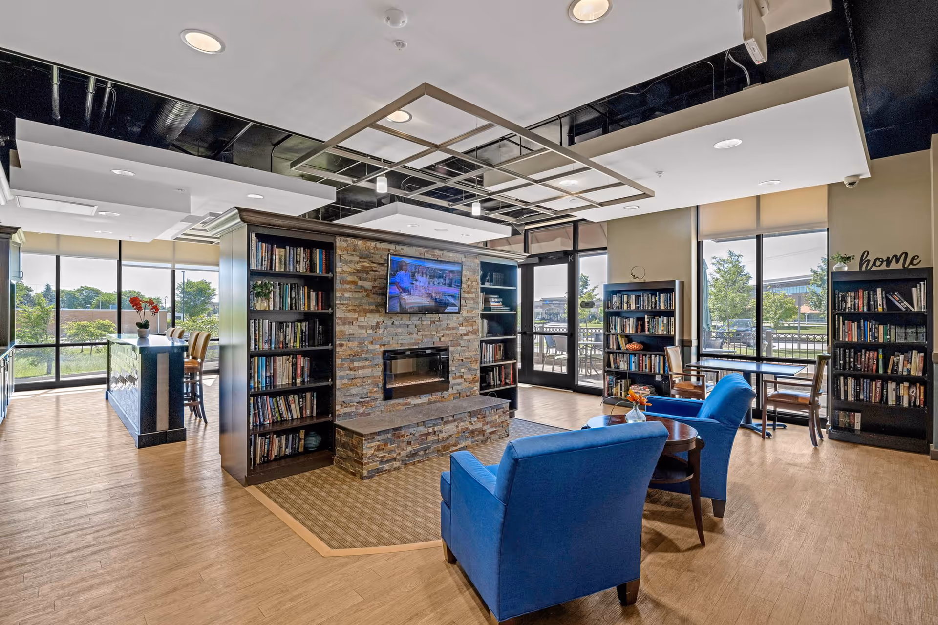 Contemporary senior living lounge with blue chairs, stone fireplace, bookshelves, and windows.