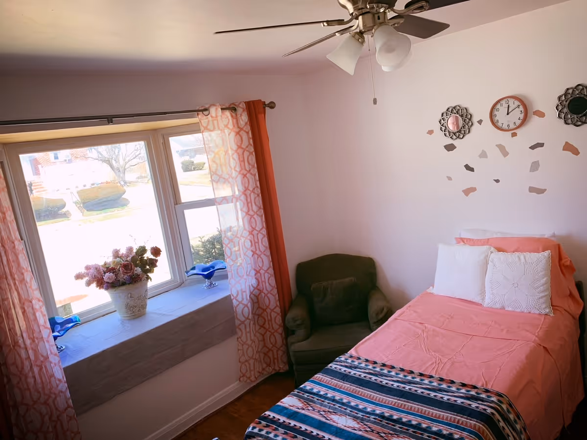 Cozy pink bedroom with bed, window seat, chair, and decorative wall clocks.