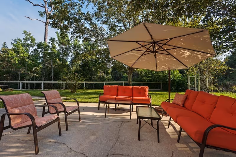 Outdoor patio with red-cushioned sofas and chairs, a large tan umbrella, and a grassy tree-lined background.